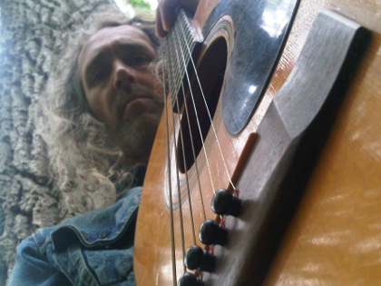 photo of Andy with acoustic guitar in foreground, trees in the background, taken in the woods behind his house in Ithaca.